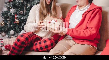 Romantisches Paar tauscht Geschenke in der Nähe geschmückten weihnachtsbaum. Mann gibt weihnachtsgeschenk Überraschung lächelnde Frau im Pullover beim Entspannen auf Co Stockfoto