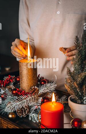 Frau Hände Blitz große goldene Kerze mit Streichhölzern in Weihnachtskranz. Weihnachten Innendekoration auf rustikalem Holzhintergrund. Hygge, Ornamente, Kerze Stockfoto