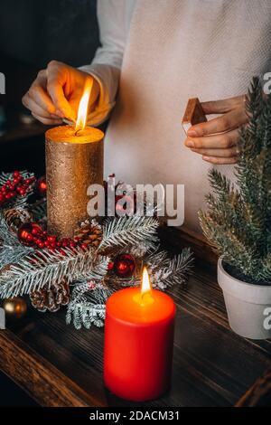 Frau Hände Blitz große goldene Kerze mit Streichhölzern in Weihnachtskranz. Weihnachten Innendekoration auf rustikalem Holzhintergrund. Hygge, Ornamente, Kerze Stockfoto