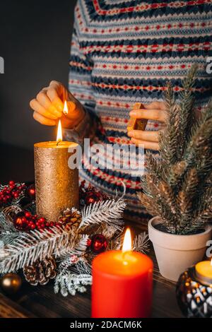 Frau Hände Blitz große goldene Kerze mit Streichhölzern in Weihnachtskranz. Weihnachten Innendekoration auf rustikalem Holzhintergrund. Hygge, Ornamente, Kerze Stockfoto