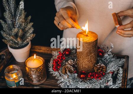 Frau Hände Blitz große goldene Kerze mit Streichhölzern in Weihnachtskranz. Weihnachten Innendekoration auf rustikalem Holzhintergrund. Hygge, Ornamente, Kerze Stockfoto