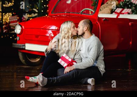 Ein junger Mann und ein junges blondes Mädchen, die sich gegenseitig lieben, umarmen und küssen sich in gemütlichen Pullovern nahe einem weihnachtlichen Hintergrund. Winter, Paar, weihnachtskonzept. Stockfoto