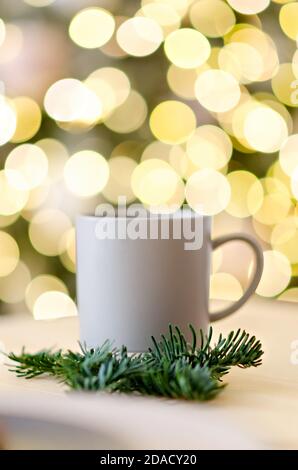 Grußkarte zum neuen Jahr. Auf dem Holztisch befindet sich eine graue Tasse heißer Schokolade, verziert mit einem Christbaumzweig. Verwackelte helle gelbe Lichter Bokeh Stockfoto