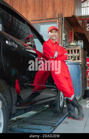 Autowaschanlage mit roter Uniform, die gegen das Auto gelehnt steht In der Waschanlage Stockfoto