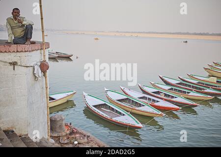 Varanasi, Indien, November 2015. Ein Mann, der in einem Ghat am Ganges mit Ruderbooten im Hintergrund sitzt. Stockfoto