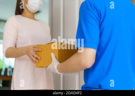 Asiatische Lieferung Mann trägt Gesichtsmaske und Handschuh in blauer Uniform hält einen Karton-Boxen im Vorderhaus und Frau eine Lieferung von Kisten von d Stockfoto