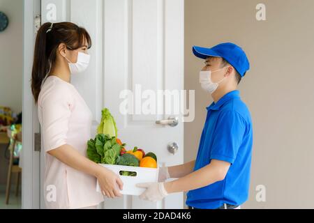 Asiatische Frau Kunde trägt Gesichtsmaske und Handschuh erhalten Lebensmittel-Box mit Lebensmitteln, Obst, Gemüse und trinken von Lieferung Mann vor dem Haus d Stockfoto