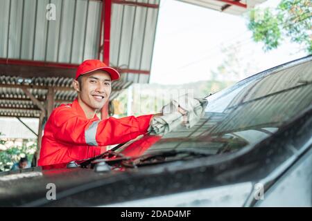 Asiatische männliche Auto Reiniger tragen rote Uniform lächelt beim Abwischen Autoglas im Autosalon Stockfoto