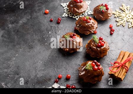 Kleine Schokoladen-Muffins werden mit Schokolade gegossen, verziert mit Winterbeeren. Zimtstangen, Schneeflocken-Dekor auf einem dunklen Tisch. Flach liegend Stockfoto