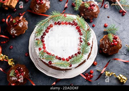 Kleine Schokoladen-Muffins und großer Kuchen werden mit Schokolade gegossen, verziert mit Winterbeeren. Zimtstangen, Schneeflocken-Dekor auf einem dunklen Tisch. Flach Stockfoto