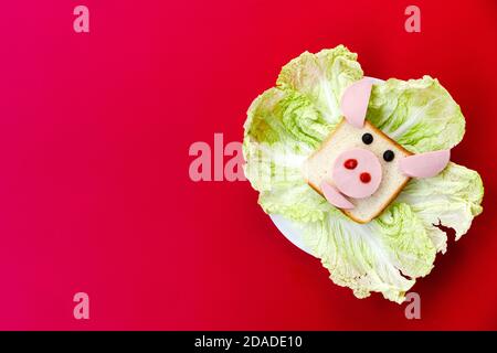 Ein Sandwich in Form eines Stierkopfes auf einem Teller, der mit Salatabgüssen verziert ist. Platte steht auf rotem Hintergrund Stockfoto