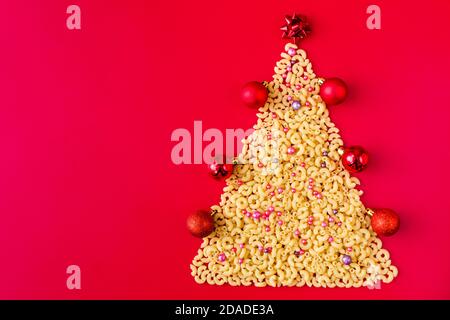 Weihnachtsbaum aus roher italienischer Pasta mit roten Kugeln isoliert auf rotem Hintergrund. Konzept für Winterferien Stockfoto