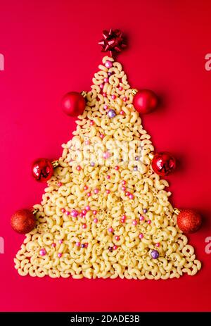 Weihnachtsbaum aus roher italienischer Pasta mit roten Kugeln isoliert auf rotem Hintergrund. Konzept für Winterferien Stockfoto