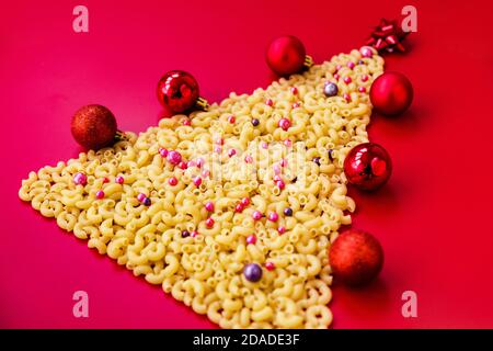 Weihnachtsbaum aus roher italienischer Pasta mit roten Kugeln isoliert auf rotem Hintergrund. Konzept für Winterferien Stockfoto