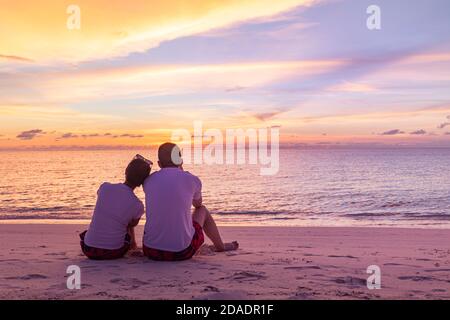 Ein Paar liebt es, den Sonnenuntergang an den Sommerferien am Strand zu beobachten. Die Silhouette der Menschen von hinten sitzend und genießen den Blick auf das Meer bei Sonnenuntergang auf tropischem Meer Stockfoto