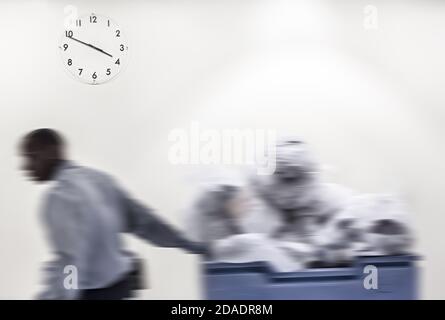 Zeitbegriff. Verschwommenes Bild des schwarzen Mannes trägt den Wagen mit Müll vor Wandhintergrund mit einer Uhr Stockfoto