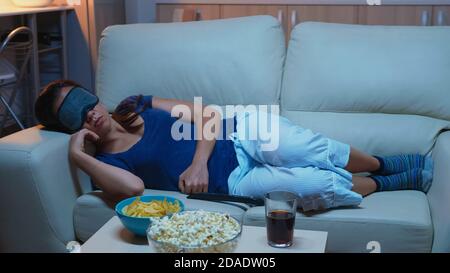 Junge Dame, die auf dem Sofa ruht und eine Maske zum Schlafen trägt, liegt auf dem gemütlichen Sofa. Müde erschöpft einsam schläfrig Frau im Schlafanzug Einschlafen auf der Couch vor dem Fernseher, Augen schließen, während Film zu sehen Stockfoto