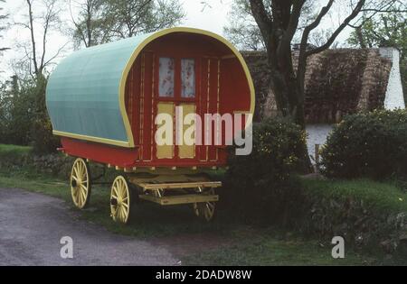 70er Jahre, traditionelle Gyspy Wohnwagen oder romany Bowtop Wagen auf einem Grasrand vor einem traditionellen Reetdachhaus verpackt. Handgebaute Holzgagone wie dieser, waren unter Romani und anderen Reisegruppen für ihre praktisch geschätzt, da sie sehr wendig und stabil waren. Stockfoto