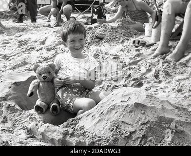 1960s, historischer, süßer kleiner Junge, der auf einem Sandstrand sitzt, mit seinem Teddybär, England, Großbritannien. Stockfoto