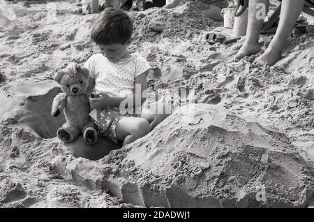 1960s, historischer, süßer kleiner Junge, der auf einem Sandstrand sitzt, mit seinem Teddybär, England, Großbritannien. Stockfoto