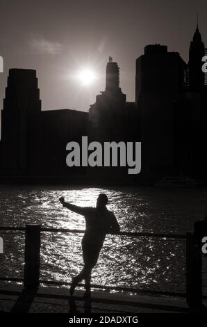 Silhouette eines jungen Mannes, der ein Selfie auf dem Hintergrund des East River und der untergehenden Sonne mit Die Wolkenkratzer von Manhattan in NYC Stockfoto