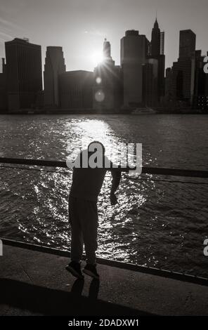 Silhouette eines Jungen, der am Ufer des Ostens steht Fluss und Blick auf die untergehende Sonne auf dem Manhattan Wolkenkratzer Hintergrund in NYC Stockfoto