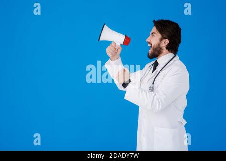 Ein junger bärtiger Arzt auf blauem Hintergrund mit einem Megaphon in seinen Händen schreit Stockfoto