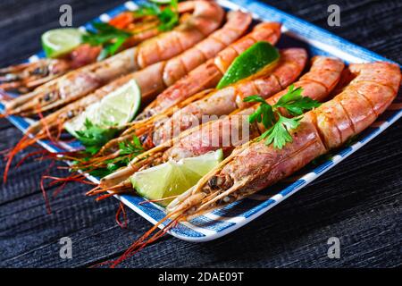 Geröstete Garnelen oder Garnelen auf einem rechteckigen Teller mit Limettenkeilen und Petersilie auf dunklem Holzgrund, Draufsicht, Nahaufnahme Stockfoto