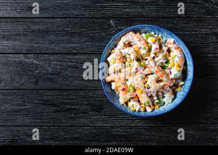 Garnelensalat mit saurem Sahne-Dressing, Mais, rote Zwiebeln und Petersilie auf einer blauen Salatschüssel mit Besteck und Crackern auf dunklem Holzhintergrund serviert, Stockfoto