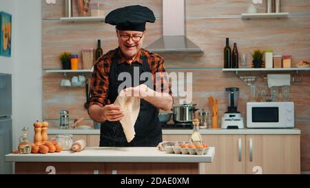 Koch wirft Teig für Pizza zu Hause in der modernen Küche schmort vor der Kamera. Geschickter älterer Koch im Ruhestand trägt Uniform Spinnen und werfen Pizza Arbeitsplatte Stockfoto