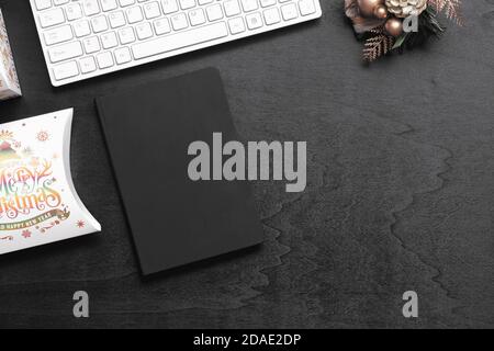 Draufsicht auf leere schwarze Abdeckung Notebook auf schwarzem Holz Home Office Schreibtisch Hintergrund mit Weihnachten Dekorationen und Weihnachten Geschenkbox. Mockup Weihnachten Backgr Stockfoto
