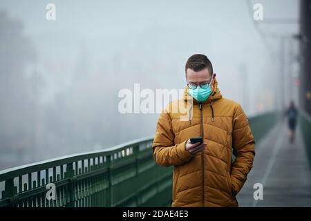 Einsamer Mann mit Gesichtsmaske mit Telefon während des Spaziergangs auf Brücke gegen die Stadt in geheimnisvollen Nebel. Düsteres Wetter in Prag, Tschechische Republik. Stockfoto