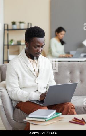 Afrikanischer Mann sitzt auf dem Sofa vor dem Tisch Mit Büchern und Tippen auf dem Laptop lernt er online bei Zu Hause Stockfoto