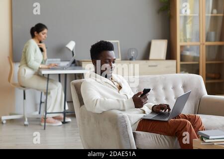 Afrikanischer Mann sitzt auf dem Sofa mit Laptop und mit Handy Telefon mit Frau, die im Hintergrund zu Hause arbeitet Stockfoto