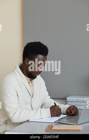 Afrikanischer Mann sitzt an seinem Arbeitsplatz mit Laptop und Schreiben Businessplan im Notizblock Stockfoto