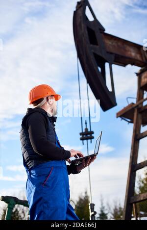 Ölmann hält Notizbuch und betrachtet die Kippmaschine der Ölpumpe. Männlicher Arbeiter in Uniform und Helm mit Laptop und Steuerung der Arbeit der Pumpe Jack. Konzept der Erdölindustrie und Ölförderung. Stockfoto