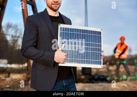 Nahaufnahme eines lächelnden Geschäftsmannes in Anzugjacke, mit Mini-Solarpanel, im Ölfeld stehend mit Daumen nach oben, Ölarbeiter vor verschwommenem Hintergrund. Konzept der alternativen Energie Stockfoto