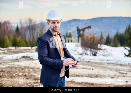 Vorderansicht des Ölmanns in Schutzhelm Schreiben auf Zwischenablage während der Arbeit im Ölfeld. Lächelnder bärtiger Gentleman in karierter Jacke, der Notizen macht. Konzept der Erdölindustrie. Stockfoto