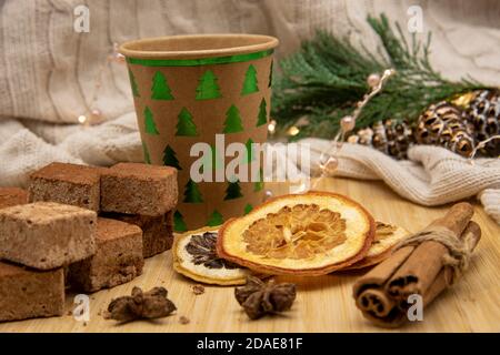 Einweg-Weihnachtsglas aus Pappe mit einem Getränk und verschiedenen Süßigkeiten und aromatischen Gewürzen auf einem Holztablett. Stockfoto