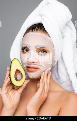 Frau mit Handtuch auf dem Kopf und Maskenblatt auf dem Gesicht Halten Avocado isoliert auf grau Stockfoto