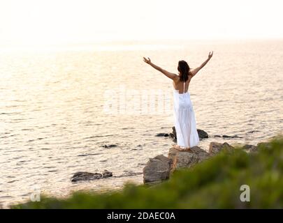 Blick von hinten auf eine nicht erkennbare Frau mit weiß lang Zieh dich an und öffne die Arme nach oben und schaust auf die untergehende Sonne - allein weibliche Person mit barfuß o Stockfoto