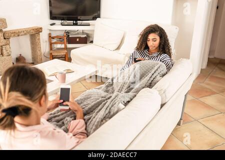 Mischrasse weibliches Paar unter einer Decke auf dem ruhen Sofa mit Smartphone, um Freunde online mit sozialen Netzwerken zu erreichen - zwei junge Frauen Techno Stockfoto