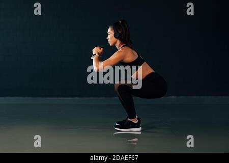 Seitenansicht der muskulösen Frau mit Kopfhörern tun hockt in Fitnessraum Stockfoto