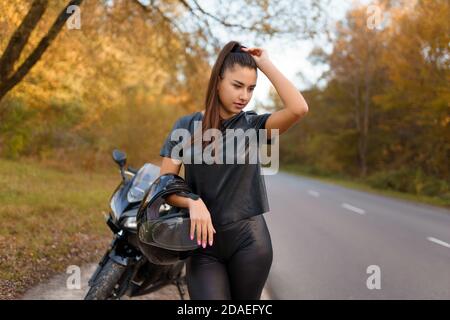 Motorradfahrerin steht in der Nähe von Motorrad am Straßenrand und hält Helm Stockfoto