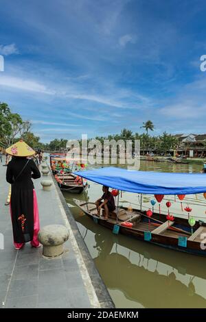 Eine Frau mit traditionellem vietnamesischem konischem Hut, eine Non La, steht auf der Bach Dang Street neben einem Boot auf dem Fluss Thu Bon, Hoi an, Vietnam, Asien Stockfoto