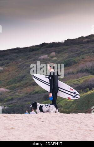 Ein Surfer mit seinem Surfbrett, der mit seinem Hund am Holywell Beach in Cornwall spazierengeht. Stockfoto