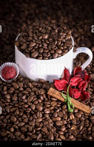 Weiße Tasse voller Kaffeebohnen auf einem Hintergrund mit roten Blumen und Zimt. Espresso am Morgen. Kaffeetasse. Stockfoto