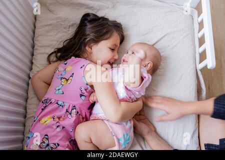 Fröhliches kleines Mädchen lächelt und umarmt ihre glückliche Baby Schwester Auf einem Bett Stockfoto