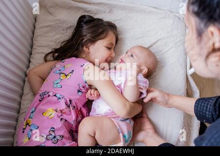 Fröhliches kleines Mädchen lächelt und umarmt ihre glückliche Baby Schwester Auf einem Bett Stockfoto