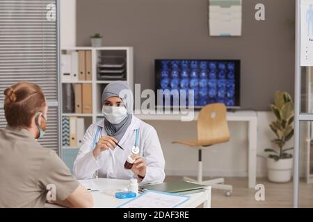 Muslimische Ärztin in Schutzmaske Verschreibung der Medizin zu Der männliche Patient, während sie im Krankenhaus sitzen Stockfoto